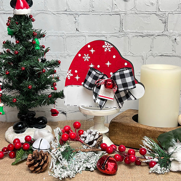 Janet's Craft Corner presents the Chunky Santa Hat, featuring a charming buffalo check bow and adorned with small evergreen branches, red berries, bells, and a mini Christmas tree against a white brick background—a perfect addition to your holiday home decor.