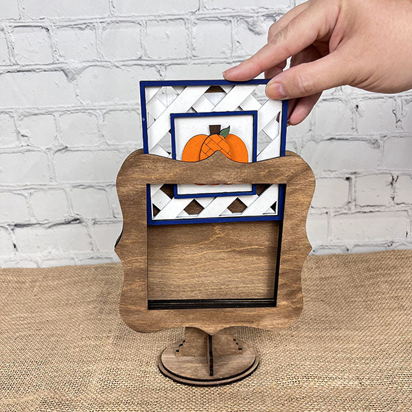 A hand is inserting an interchangeable square sign featuring a pumpkin design into Janet's Craft Corner's Drop In Interchangeable Squares Holder, which has a stylish wooden frame. The backdrop is a white brick wall with the sign resting on a textured mat, ideal for showcasing your creativity like a DIY kit or as a drop-in sign display.