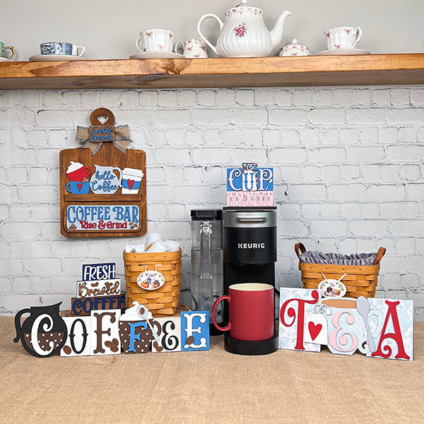 A caffeine enthusiast’s coffee bar has a Keurig, red mug, baskets with cups and supplies, decorative signs, teapots, and the Janet's Craft Corner Coffee Word Block DIY craft kit set on a wooden shelf against a white brick wall.