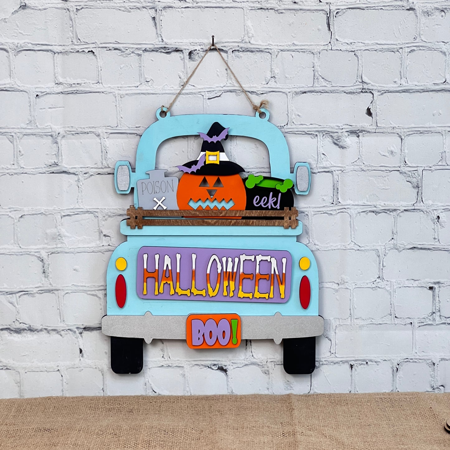 Janet's Craft Corner offers a charming Halloween decor DIY kit, featuring a light blue vintage truck with "HALLOWEEN" and "BOO!" in lively colors. It includes a pumpkin insert, witch's hat, broom, and playful words like "poison" and "eek!" for a festive touch.