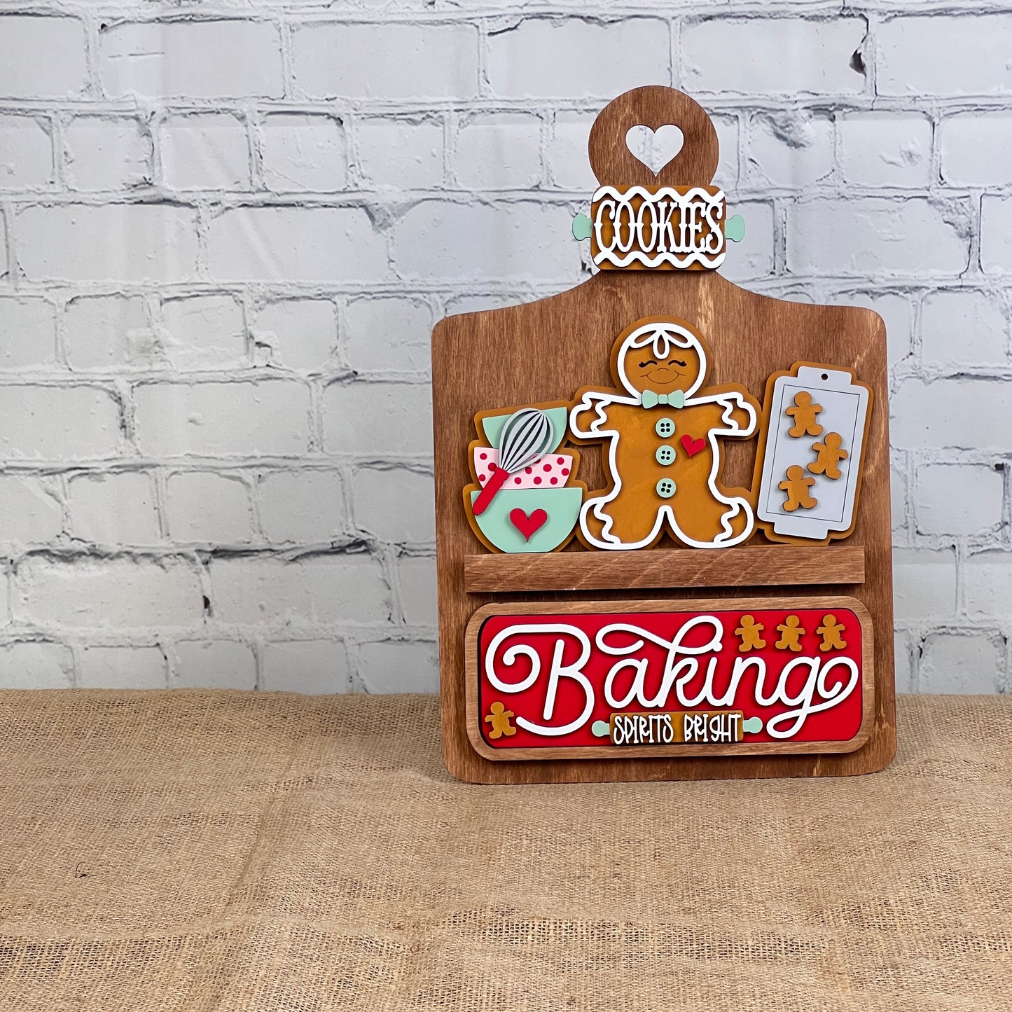 A wooden stand set against a brick wall displays gingerbread-themed decorations, featuring cookie-shaped elements and a "Baking Spirits Bright" interchangeable insert from Janet's Craft Corner. This delightful arrangement, topped with a "Cookies" sign, offers the perfect opportunity for personalization to create an ideal DIY craft kit experience.