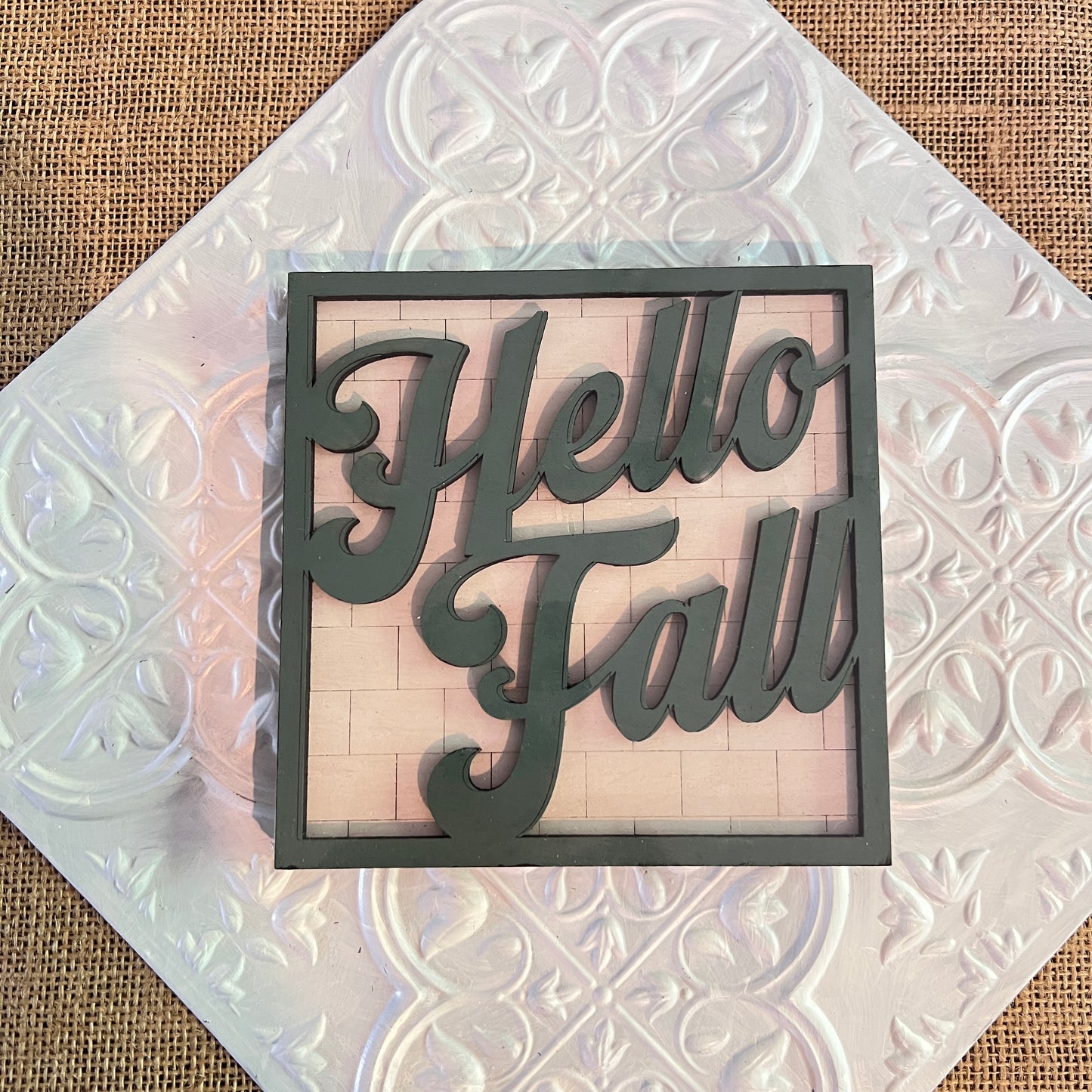 A decorative "Hello Fall" sign in cursive rests on a 6 inch square from Janet's Craft Corner atop a burlap surface.