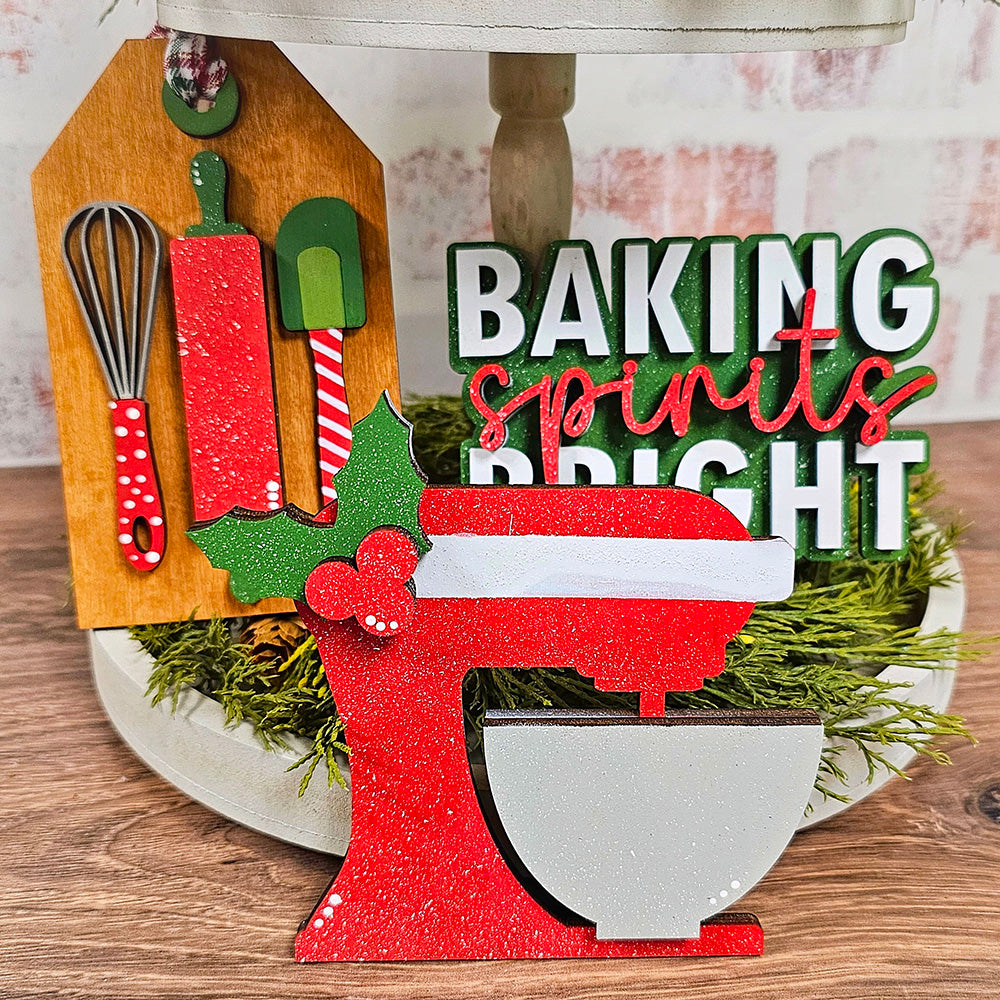 The Baking Spirits Bright Decor Set by Janet's Craft Corner adds holiday cheer to your festive kitchen with a red stand mixer adorned with holly, complemented by a delightful sign that reads "Baking Spirits Bright." It also includes a wooden plaque holding a whisk, spatula, and cutting board. Everything is beautifully arranged on a wood surface embellished with greenery.
