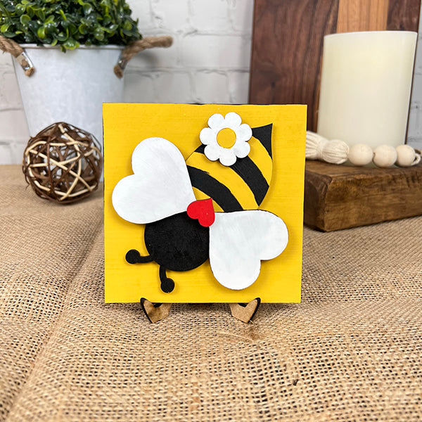 The Bee Swappable Squares by Janet's Craft Corner features a handmade bee with white wings, yellow and black body, white flower, and red heart on a wooden stand atop burlap—ideal for adding rustic charm to your home decor.