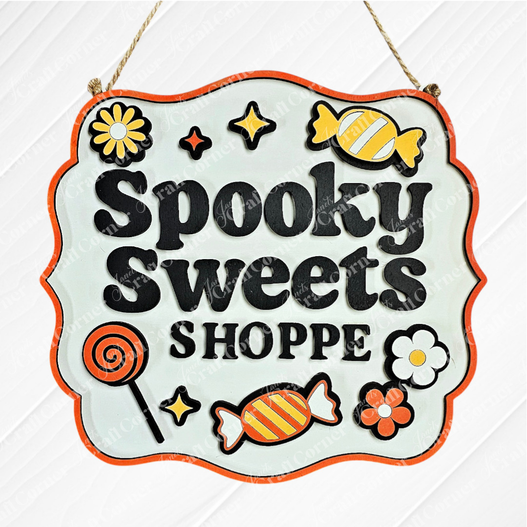 The Spooky Sweets Shoppe Wall Hanger by Janet's Craft Corner features bold lettering with candies, flowers, a lollipop, and stars on white with an orange border—ideal for seasonal decor or as a DIY project.