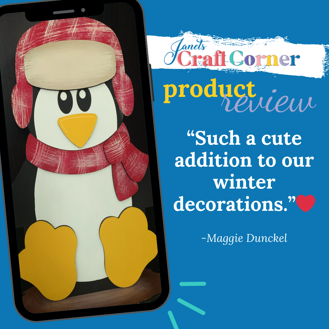 A smartphone displays the Woodland Friends Penguin Shelf Sitter from Janet's Craft Corner, adorned with a red hat and scarf. Text reads: "Janet's Craft Corner product review" featuring Maggie Dunckel's quote: “Such a cute addition to our winter decorations.” A red heart adds warmth.