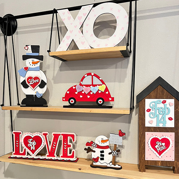 Wooden shelves feature Janet's Craft Corner's Love Shelf Sitter DIY kit, offering Valentine's Day home décor with "XO," "LOVE," heart-holding snowmen, a red car with hearts, and a house shape detailing signs like "Feb 14" and "Be Mine.