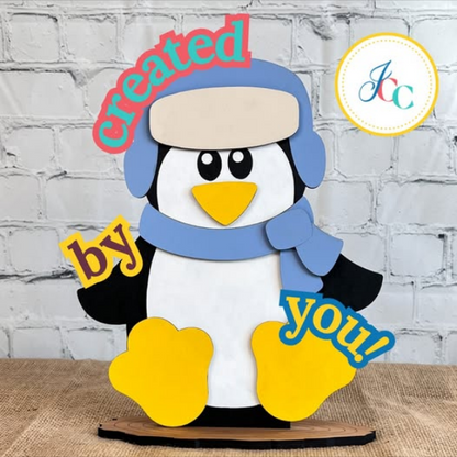 The Woodland Friends Penguin Shelf Sitter by Janet's Craft Corner features a cartoon penguin in a blue hat and scarf, yellow feet, leaning against a brick wall. "Created by you!" appears in colorful letters with the "Jcc" logo encouraging creativity.