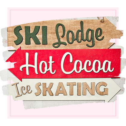 The Ski Lodge Directory Sign by Janet's Craft Corner features three arrows: "Ski Lodge" in green, "Hot Cocoa" in white on red, and "Ice Skating" in brown. This DIY seasonal home decor craft kit adds charm to any home.