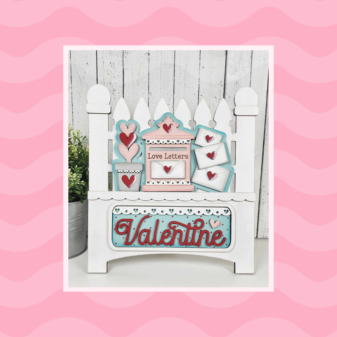 Janet's Craft Corner offers a DIY home decor kit featuring a Garden Fence Interchangeable Base with an insert displaying heart motifs, "Love Letters," and "Valentine" in red on a pink wavy background, completed by a potted plant for a charming Valentine's display.