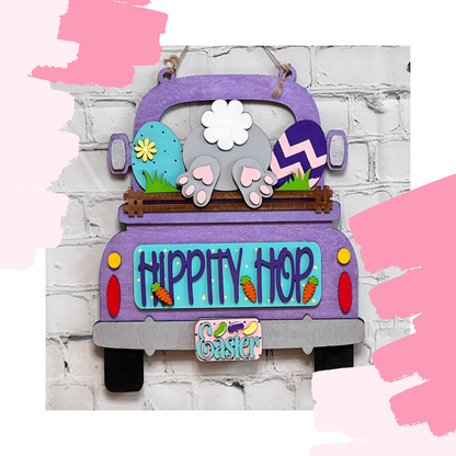 Janet's Craft Corner offers a DIY home decor kit featuring an Antique Truck Interchangeable Base with a purple truck insert. The design includes a bunny's back and feet, Easter eggs, "Hippity Hop," painted carrots, and the "Easter" license plate. It's ideal for interchangeable seasonal decor.