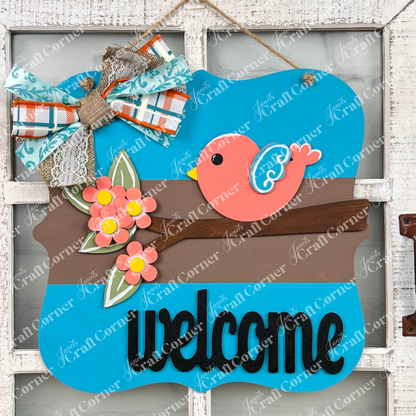 The Bird Welcome Sign by Janet's Craft Corner features a vibrant pink bird with blue wings perched on a brown branch, highlighted by delicate pink flowers. A stylish bow interweaving floral and plaid designs adds a decorative touch to the corner. Ideal for home décor, it elegantly displays the word "welcome" in black at the bottom.