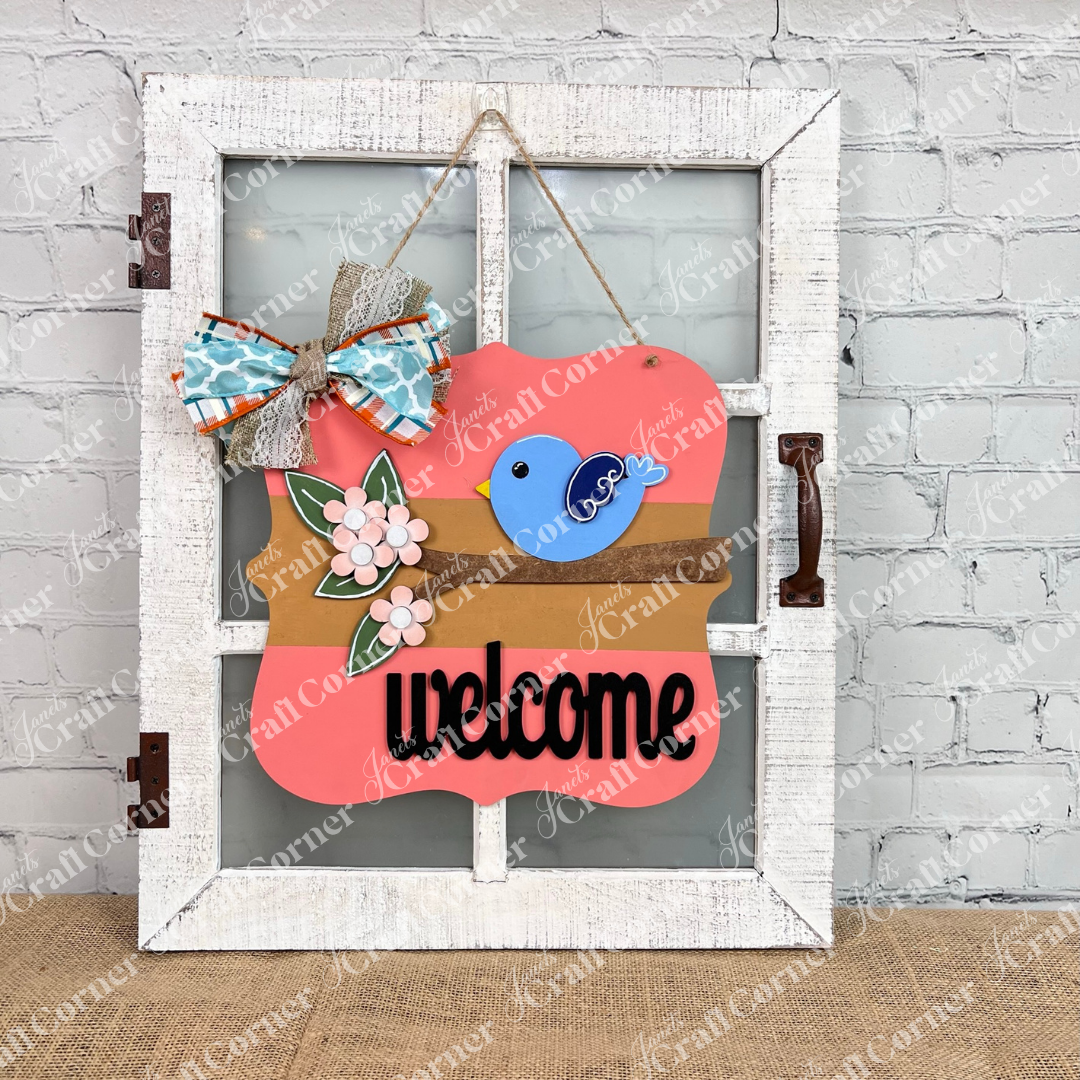 A delightful Bird Welcome Sign from Janet's Craft Corner enhances the door, featuring a blue bird on a branch decorated with pink flowers and green leaves. A vibrant bow in blue, orange, and lace patterns tops this charming home décor piece, with "welcome" prominently displayed underneath.
