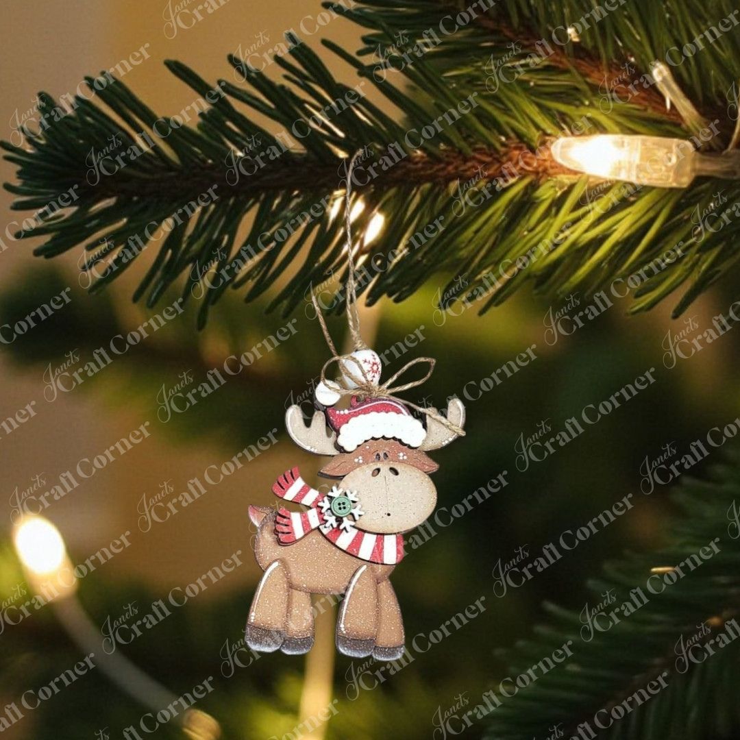 A cute reindeer from Janet's Craft Corner Forest Friends Ornaments DIY craft kits (choice of 5) wears a red scarf and Santa hat as it hangs from a Christmas tree branch, surrounded by glowing string lights.