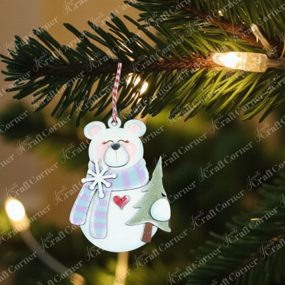 A bear-shaped ornament from Janet's Craft Corner's Forest Friends Ornaments DIY kit, featuring a scarf, heart, and tree, hangs on a pine branch with glowing white lights—one of 5 designs to craft yourself.