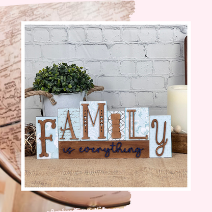 The "Family is Everything" word block shelf sitter from Janet's Craft Corner features blocks spelling "FAMILY" with the phrase beneath. It's a DIY home decor craft kit, enhanced by a potted plant and candle against a white brick wall.