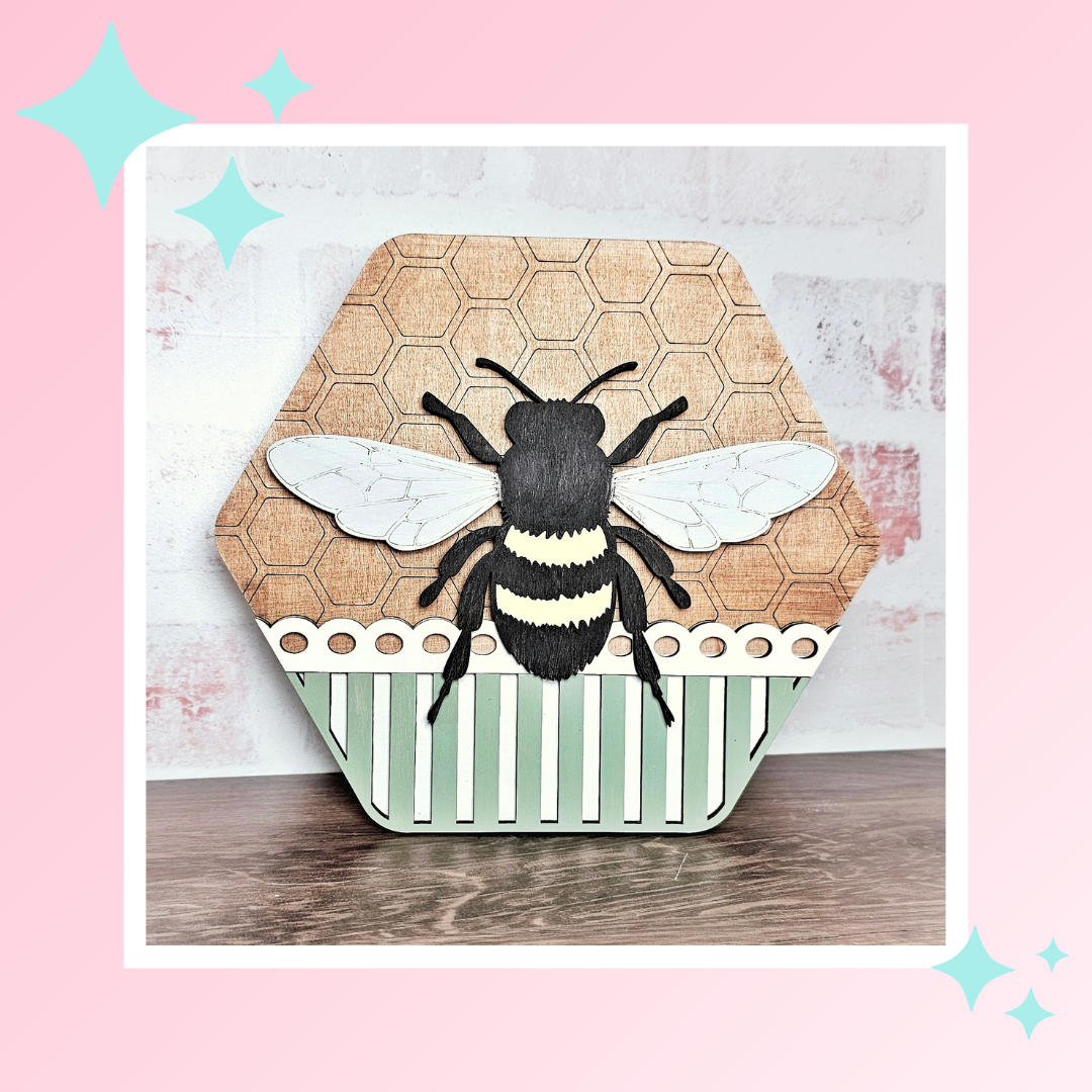 Janet's Craft Corner offers a Honeycomb Shaped Bee Sign Door from their DIY seasonal home decor craft kit. It features a bee illustration with white wings, a honeycomb pattern, green and white striped lower half with scalloped edge, and a pink background that sparkles with starry charm.