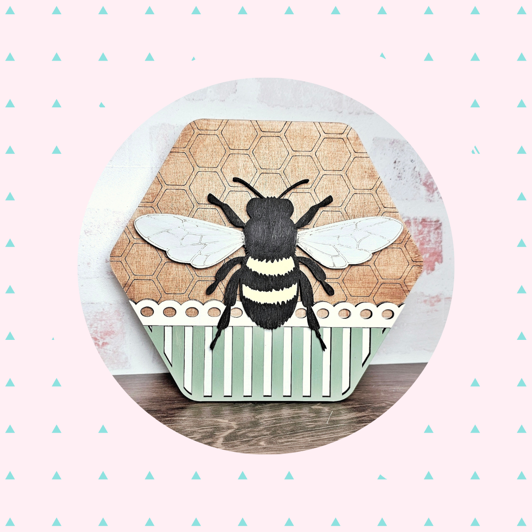 This Honeycomb Shaped Bee Sign Door kit by Janet's Craft Corner features a bee on a hexagonal wooden panel with honeycomb, white wings, and green stripes over the pink background with turquoise triangles. Ideal for seasonal DIY decor.