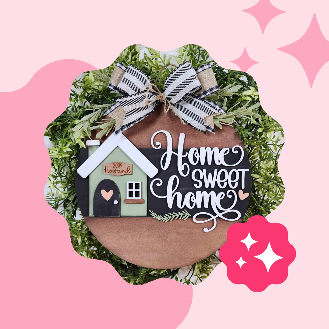 The Janet's Craft Corner "Home Sweet Home Round Door Hanger" is a DIY décor kit featuring a hand-painted small house and heart with "Home Sweet Home" on a wooden base, embellished with a checkered bow and greenery against a pink background with star accents.