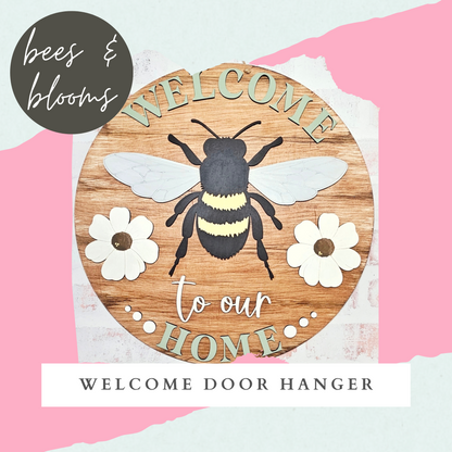 The Welcome Home Bees & Blooms Sign from Janet's Craft Corner is a charming DIY decor kit featuring a round wooden hanger with a painted bee, two white blooms, and "WELCOME to our HOME" on a pink and white abstract background, accented by the Bees & Blooms logo.