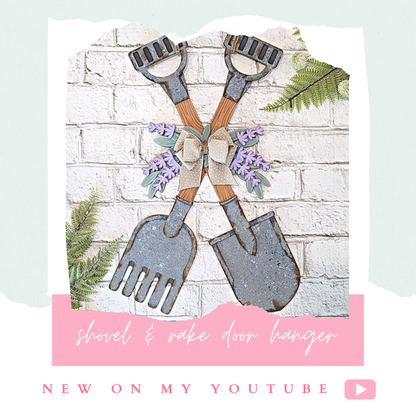 A lavender-accented wood decor with crossed shovel and rake, adorned with a bow on a white brick wall. Fern leaves surround this DIY project featuring "Shovel and Rake Door Hanger" and "NEW ON MY YOUTUBE" text next to a play button icon. Check out Janet's Craft Corner for this seasonal craft kit!.