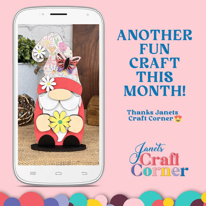 A smartphone displays the Spring Mini Shelf Sitters DIY kit from Janet's Craft Corner, featuring a decorative gnome with floral decorations and a butterfly on its hat against a pink background. Text reads, "Another fun craft this month! Thanks Janets Craft Corner." A colorful logo sits at the bottom right.