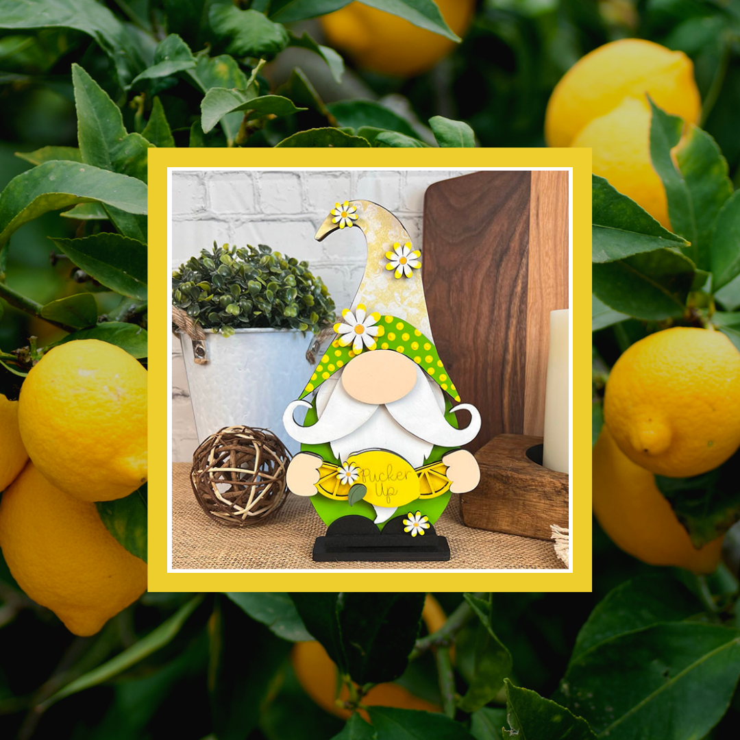 The Lemonade Gnome Shelf Sitter DIY craft kit from Janet's Craft Corner features a whimsical gnome with a green hat holding a lemon on a wooden surface, surrounded by potted plants and farmhouse decor, framed by vibrant lemons and green leaves, bringing charm to your cozy kitchen.