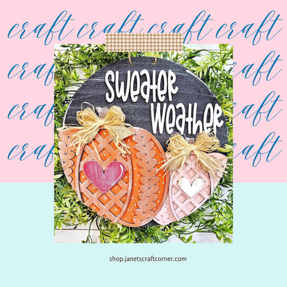 The Sweater Pumpkin Door Hanger from Janet's Craft Corner features two woven-style pumpkins, one orange and one pink, with heart cutouts and bows. It showcases "Sweater Weather" lettering against lush green foliage in a cursive pattern, making it an ideal DIY seasonal home decor craft kit.