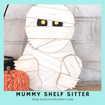 A mummy figure with overlapping white strips and big black-yellow eyes sits next to a small orange pumpkin. Text reads: "Mummy Shelf Sitter" from Janet's Craft Corner at shop.janetscraftcorner.com.