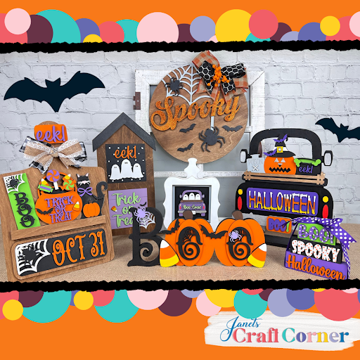 A colorful Halloween display features signs, pumpkins, bats, and Janet's Craft Corner Spooky Halloween Wall Hanger DIY craft kit—seasonal home decor with “Trick or Treat”—arranged festively on a table against a brick wall.