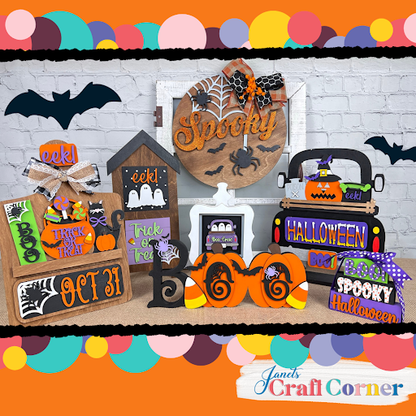 A colorful Halloween display features signs, pumpkins, bats, and Janet's Craft Corner Spooky Halloween Wall Hanger DIY craft kit—seasonal home decor with “Trick or Treat”—arranged festively on a table against a brick wall.
