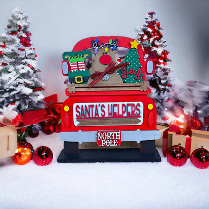 A cheerful holiday scene features a red truck with Christmas decorations like a reindeer, gifts, and a "Santa's Helpers" sign. Snowy backdrops with decorated trees and presents inspire creativity in Janet's Craft Corner's Santa’s Helpers DIY decor kit.