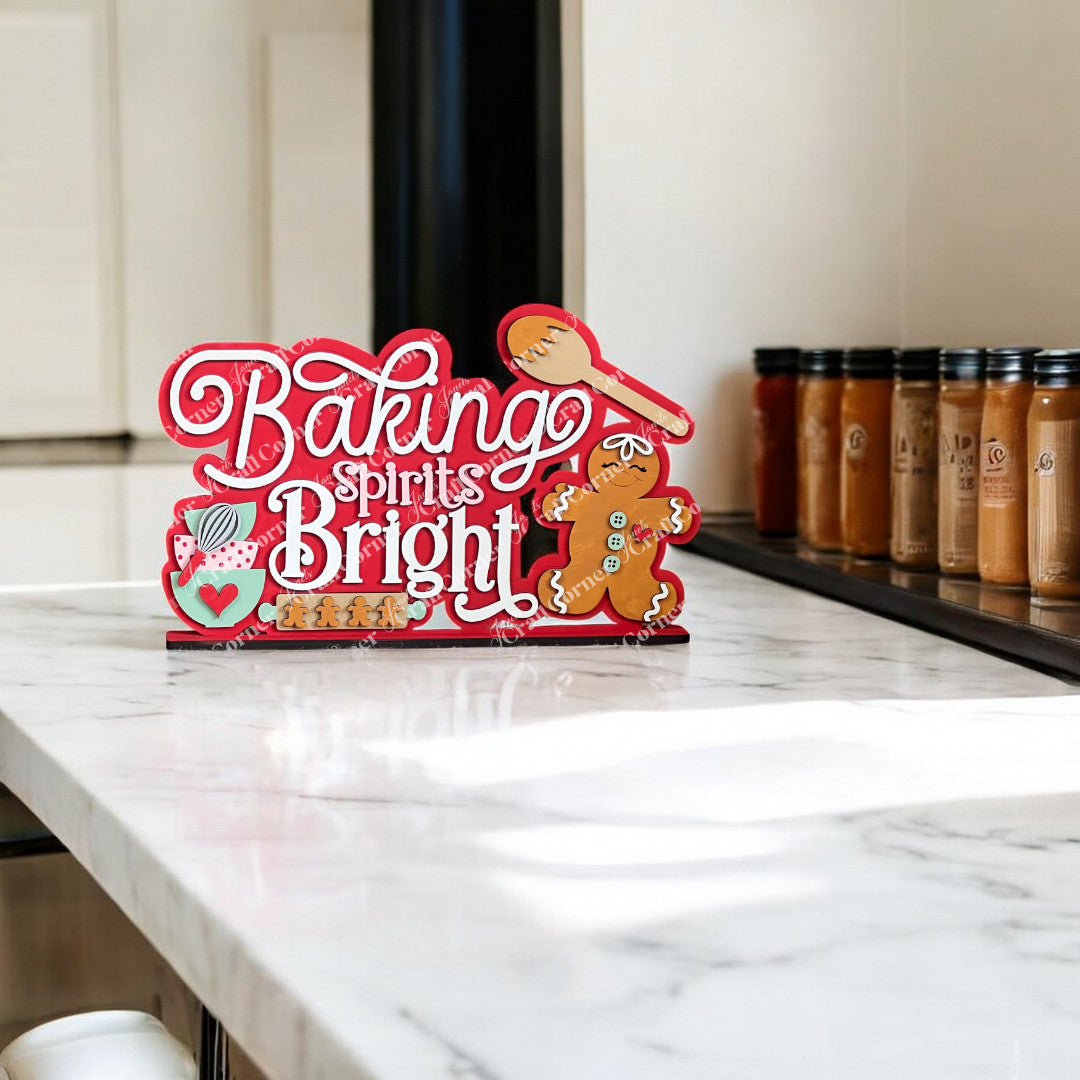 A "Baking Spirits Bright" sign sits on a marble counter, featuring festive red and white lettering with hand-painted gingerbread and kitchen motifs. The perfect piece of DIY decor from Janet's Craft Corner, it complements spice jars lining the background.