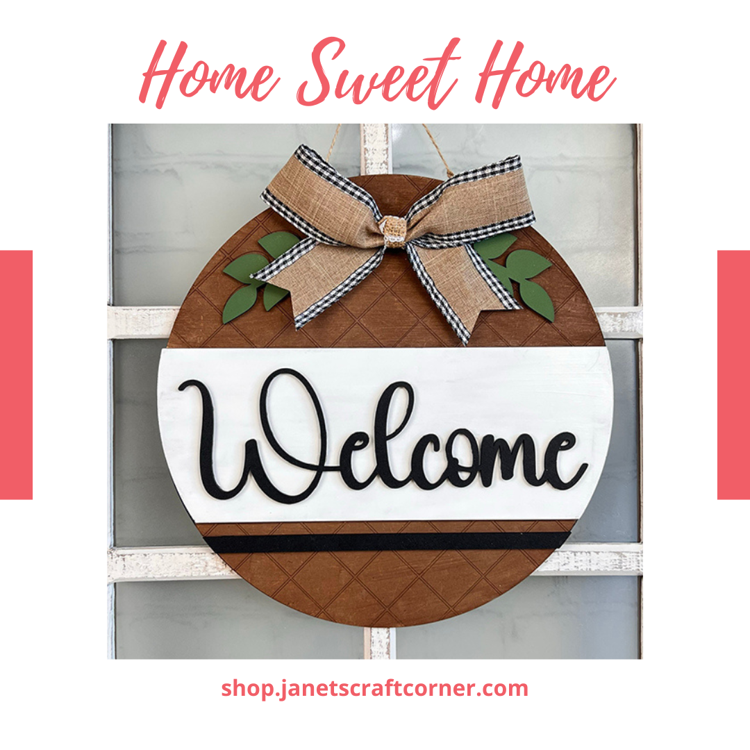 The Welcome Round Door Hanger by Janet's Craft Corner is a farmhouse decor piece with "Welcome" and "Home Sweet Home" text, adorned with a bow and leaves. It hangs on a grid and is also available as a DIY home decor craft kit.