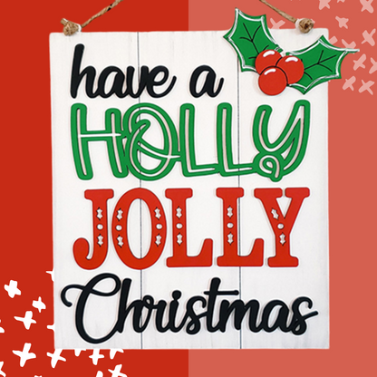 The Holly Jolly Christmas Sign from Janet's Craft Corner is a DIY home decor craft kit featuring the phrase "Have a Holly Jolly Christmas" with "holly" in green, "jolly" in red and white, holly leaves and berries in the top right, all set against a red and white background with small white Xs.