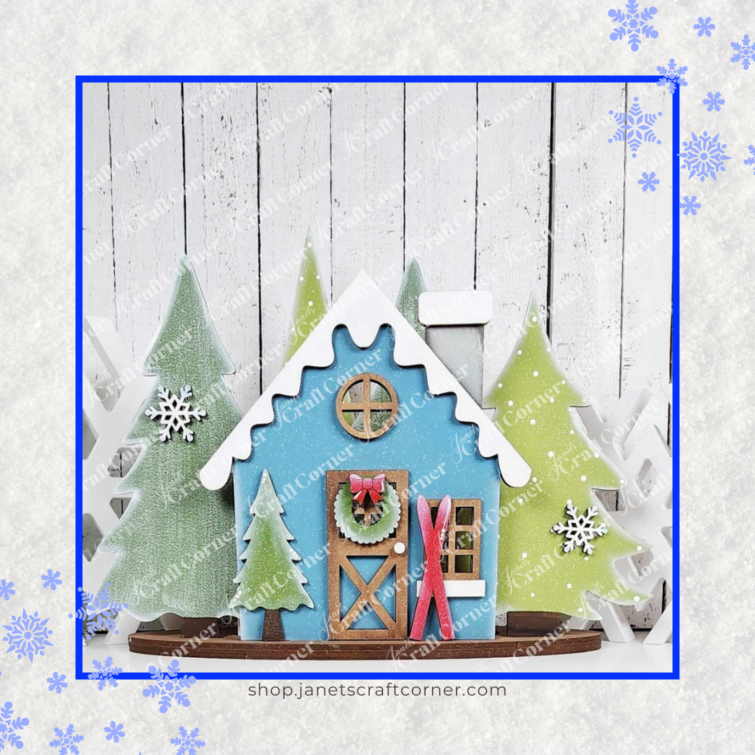 A festive scene features a blue wooden Winter Cabin Votive Holder Shelf Sitter from Janet's Craft Corner, with a snow-topped roof and wreath-adorned door, surrounded by green and white Christmas trees. Snowflakes embellish the image on a textured white wooden backdrop, ideal for cozy home decor craft kits.