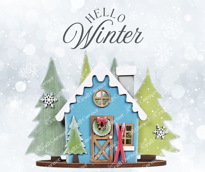 A festive winter scene features a blue house with a wreath and red skis by the door, surrounded by snowy pines and swirling snowflakes. The "Hello Winter" text above adds charm, perfect for Janet's Craft Corner DIY Winter Cabin Votive Holder Shelf Sitter as seasonal home decor.