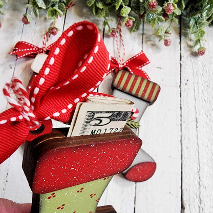 Janet's Craft Corner offers a festive Stocking Money Holder Ornament DIY kit. The wooden stocking, accented with red bows and ribbons on a white background, holds a five-dollar bill amidst greenery and red berries, adding holiday cash cheer.