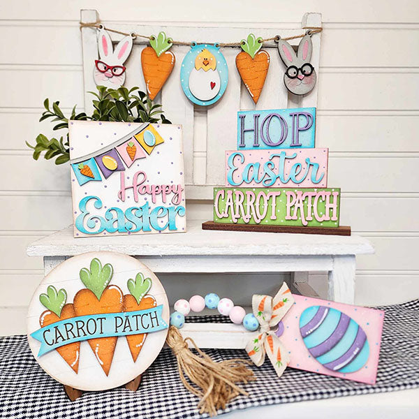 A delightful Easter display features Janet's Craft Corner Easter Tiered Tray, adorned with hand-painted signs reading "Happy Easter," "Hop," and "Carrot Patch." This enchanting decor is complemented by hanging carrots, bunny faces, and vibrant eggs, all elegantly arranged on a white bench covered in a black-and-white checkered cloth.