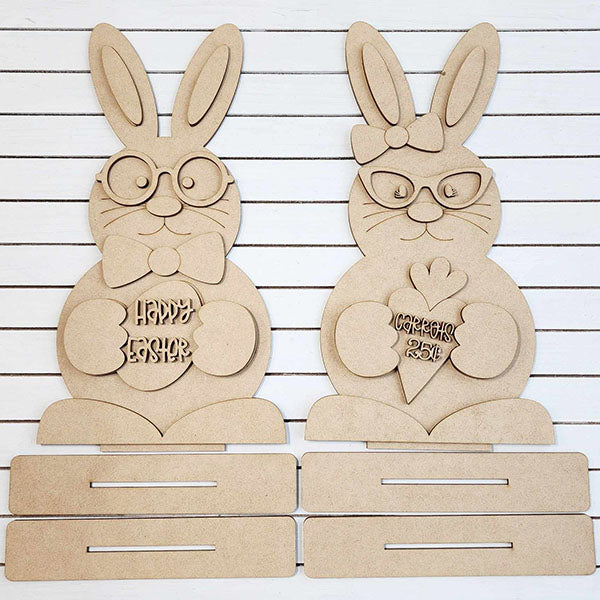 Janet's Craft Corner offers a DIY seasonal home decor kit featuring the Bunny Couple Shelf Sitters. These charming wooden Easter bunnies include one with glasses and a bowtie holding a "Happy Easter" egg, and another with glasses holding a carrot-shaped "Feliz Páscoa" piece against a slatted background.