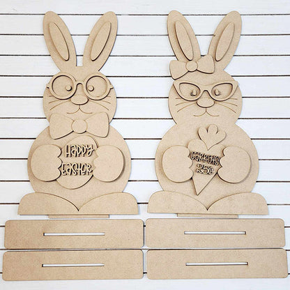 Janet's Craft Corner offers a DIY seasonal home decor kit featuring the Bunny Couple Shelf Sitters. These charming wooden Easter bunnies include one with glasses and a bowtie holding a "Happy Easter" egg, and another with glasses holding a carrot-shaped "Feliz Páscoa" piece against a slatted background.