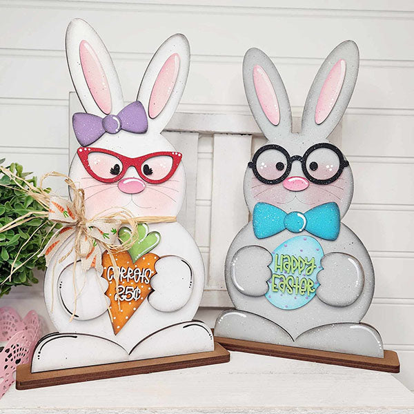 Janet's Craft Corner presents the delightful Bunny Couple Shelf Sitters, which feature two charming figures wearing glasses. One bunny holds a carrot sign inscribed with "SPRING 25¢," and the other displays an egg sign that says "HAPPY EASTER." Their adorable expressions and pastel accents provide a lovely contrast against a light background, adding cheer to your home decor.