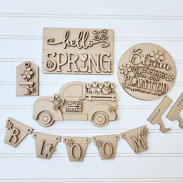 The Hello Spring Decor Set by Janet's Craft Corner is a DIY seasonal home decor craft kit featuring 6 wooden pieces: signs with "hello SPRING" and "Bloom where you are planted," a flower market truck, a small house, and a "BLOOM" banner with delicate flower details.