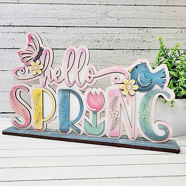 Introducing the Hello Spring Standing Shelf Sitter by Janet's Craft Corner. This delightful DIY kit features a decorative wooden sign with "hello SPRING" in pastel colors, complete with charming butterfly and bird accents perched on the letters and adorned with flower designs. Perfect for home décor enthusiasts, it makes an eye-catching display on a white wooden surface next to a green potted plant.