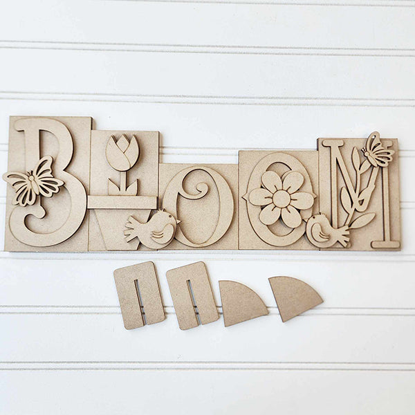 Introducing the "Bloom Word Block Shelf Sitter" from Janet's Craft Corner. This DIY home decor kit features a wood sign with carved letters: butterfly on "B," tulip on "L," bird on "O," flower on the second "O," and leaves with a bird on "M." Easy to assemble for seasonal decor.