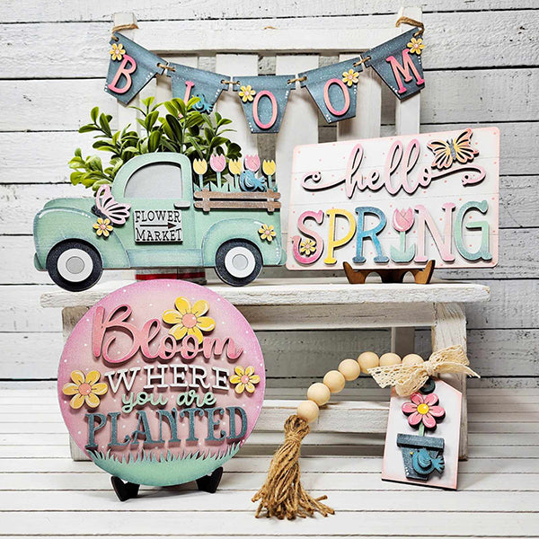 The decorative spring-themed setup from Janet's Craft Corner, featuring the "Hello Spring Tiered Tray Set," includes a charming miniature truck, signs adorned with messages like "hello SPRING" and "Bloom where you are planted," floral illustrations, and a quirky bead garland shelf sitter, all in soothing pastel colors elegantly displayed on a white bench.