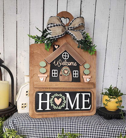 The Welcome Home Interchangeable Insert from Janet's Craft Corner is a DIY home decor kit featuring a house design with a "Welcome" message, adorned with an elegant plaid bow and greenery. The bold "HOME" letters are highlighted with a heart and wreath symbol, set against a rustic backdrop for versatile home decoration.