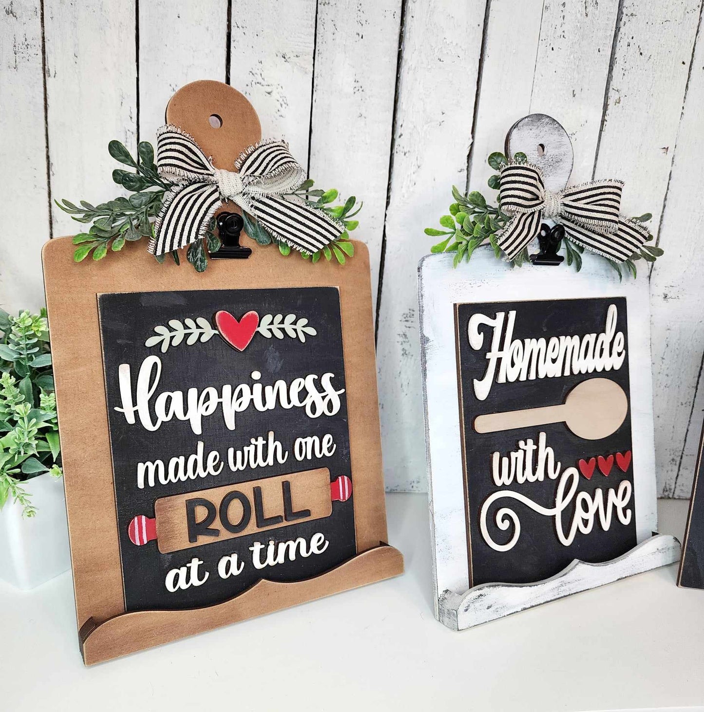 Janet's Craft Corner offers a Breadboard Sign Holder for Interchangeable Signs, perfect for farmhouse-inspired decor. This DIY home decor craft kit includes two charming designs: one reads "Happiness made with one roll at a time" featuring a rolling pin, and the other says "Homemade with Love" adorned with a spoon. Both signs are complemented by ribbon bows and greenery against a white wooden backdrop.