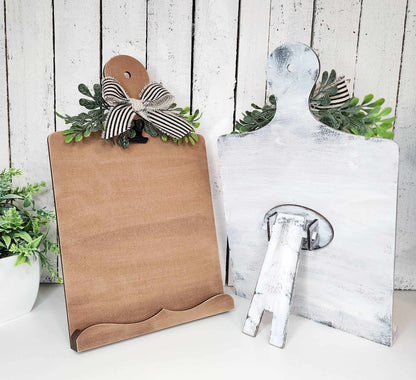 Two Breadboard Sign Holders from Janet's Craft Corner, one in a plain brown finish and the other whitewashed, each featuring leafy embellishments and a striped bow at the top, are displayed against a rustic white wooden backdrop. Perfect for farmhouse decor enthusiasts or as part of your DIY home decor craft kit.