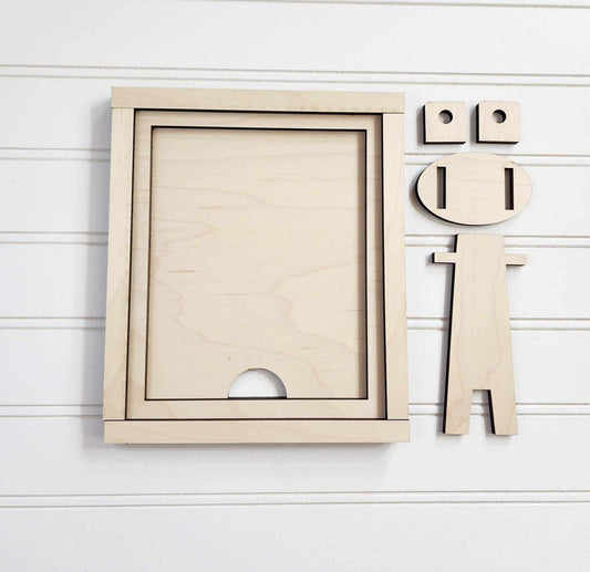 A wooden geometric figure resembling a person is positioned beside the Janet's Craft Corner Farmhouse Frame for Interchangeable Signs, which features two smaller circles on top. Set against a white, horizontally-lined background, this perfect addition to farmhouse decor has a round head with notches for eyes and mouth.